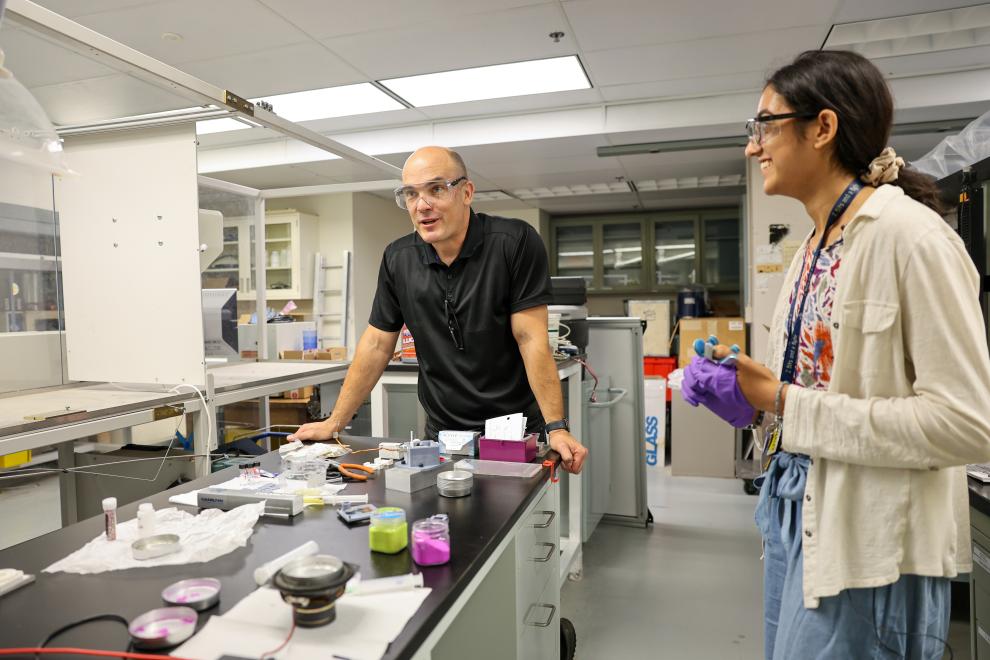 Faculty mentor in black shirt and safety glasses leaning on a lab table beside student in floral shirt and safety glasses, removing purple gloves