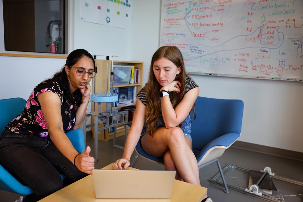 Female faculty mentor with brown hair and pink and black floral shirt sitting with student with long light brown hair and grey short sleeved shirt, talking and pointing to laptop