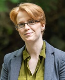 Person with short reddish blonde hair and glasses wearing a green top and grey blazer